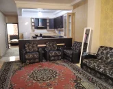اتاق پذیرایی فرش شده به همراه مبلمان تیره و آشپزخانه آپارتمان در رضوانشهر 4564634
